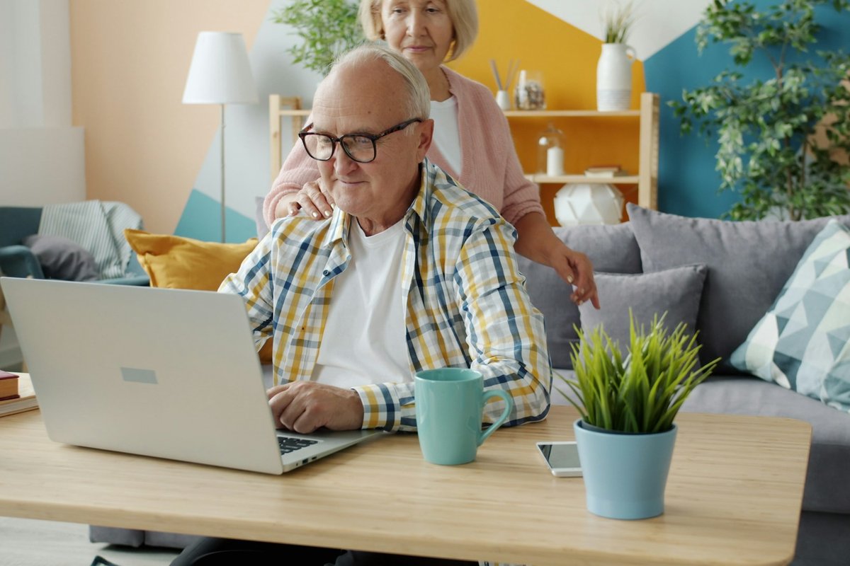 An older man using a laptop at his living room table while an older woman stands beside him with a hand on his shoulder