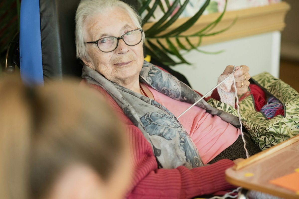 An older woman in a wheelchair at home, wearing glasses and knitting, looking up at someone sitting with her