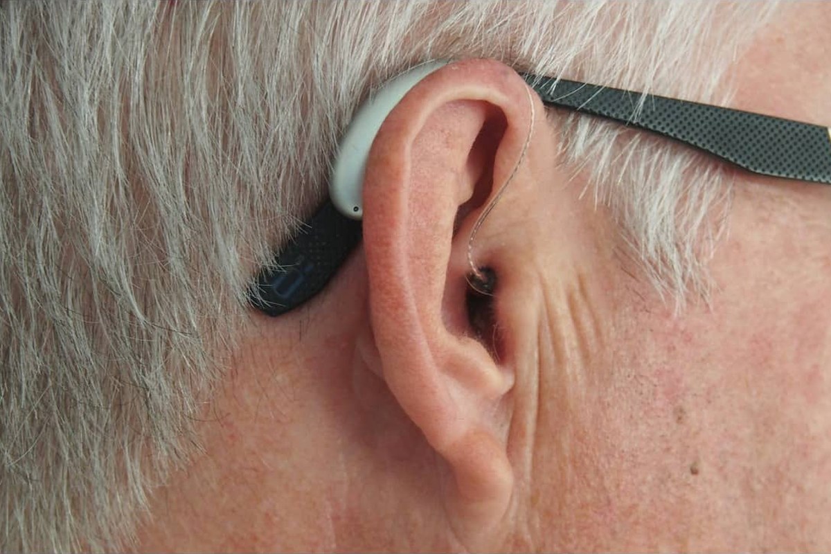 A behind-the-ear hearing aid worn by an older man with grey hair and glasses, seen in close-up