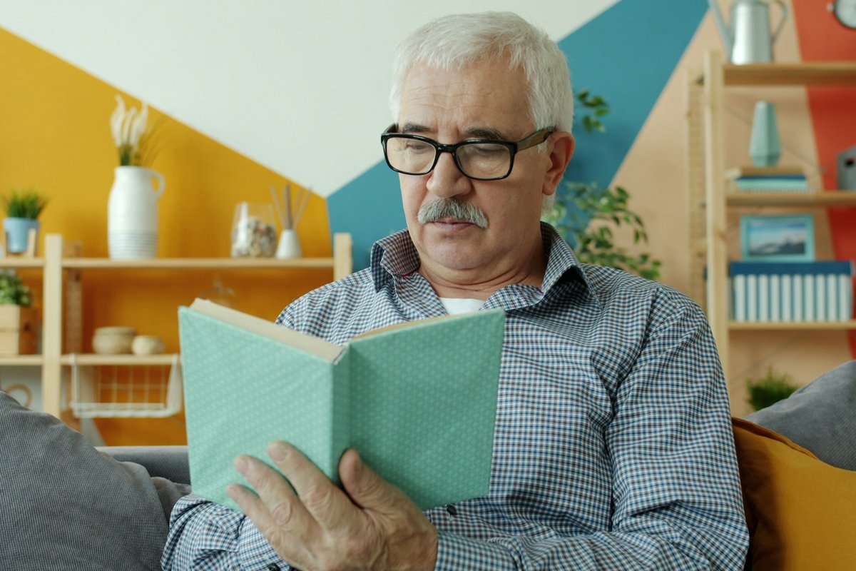 An older man wearing glasses, reading a book on a sofa in a bright living room